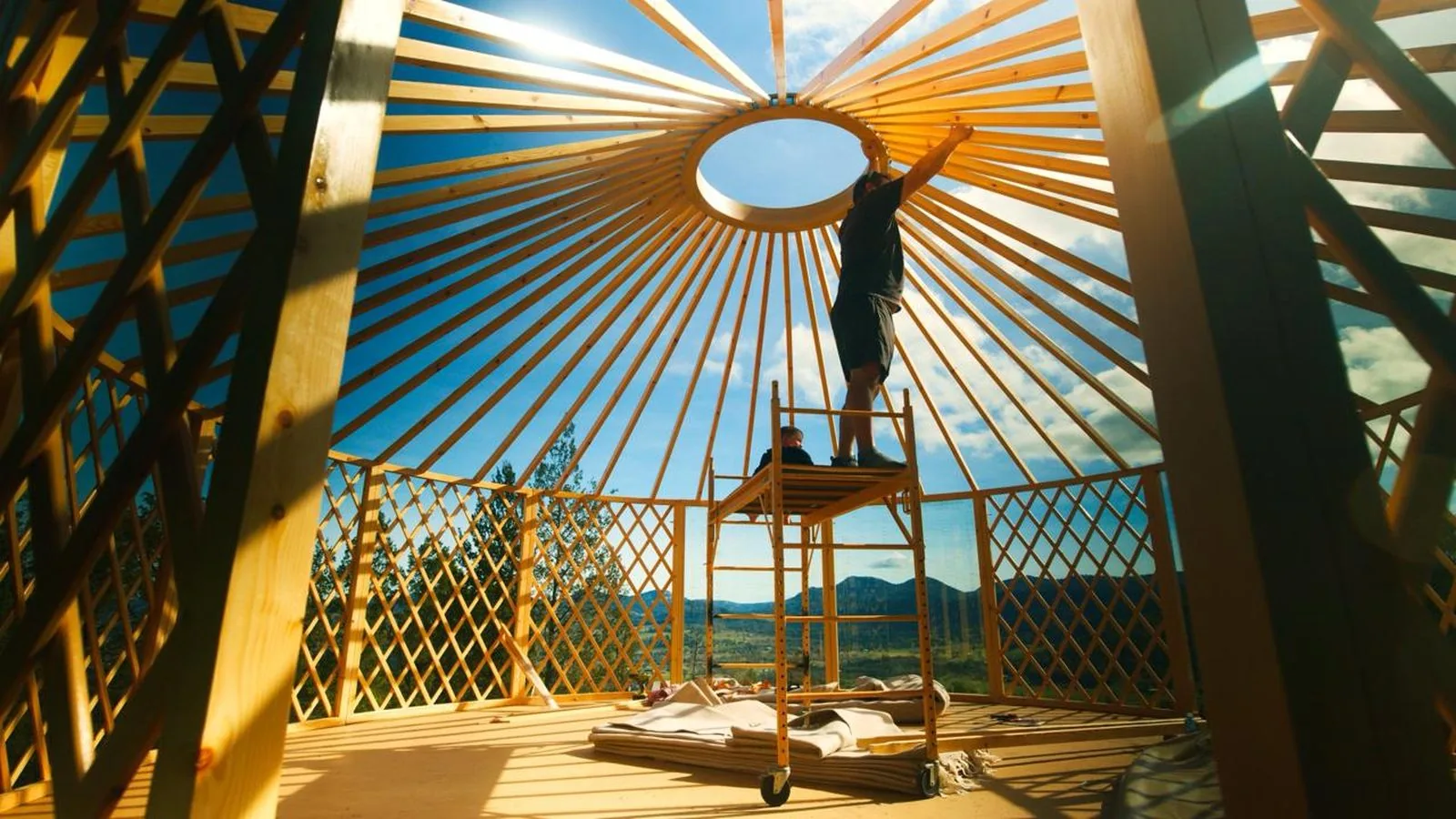 An ORON frame going up, with a builder on scaffolding fitting the crown ring and mountains visible through the lattice walls